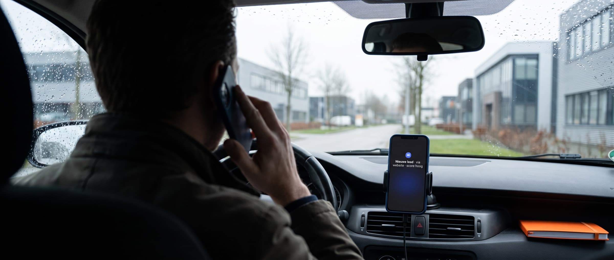 Accountmanager van een B2B-bureau belt vanuit een geparkeerde auto, met op de telefoon in de houder een melding van een nieuwe lead