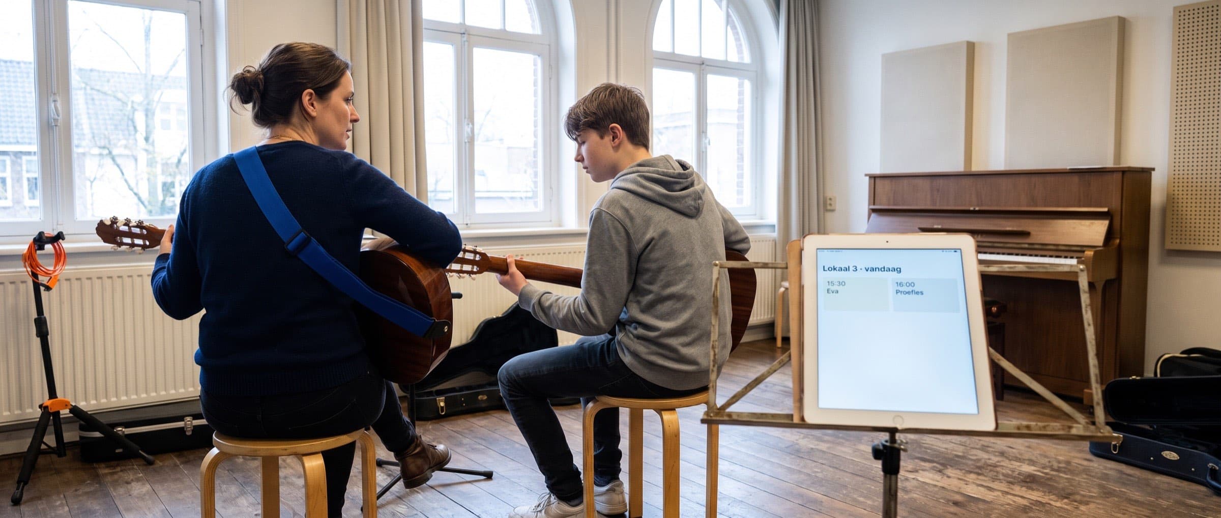 Docent en leerling tijdens een gitaarles in een lesruimte van een muziekschool, met een tablet met het dagrooster op de muziekstandaard